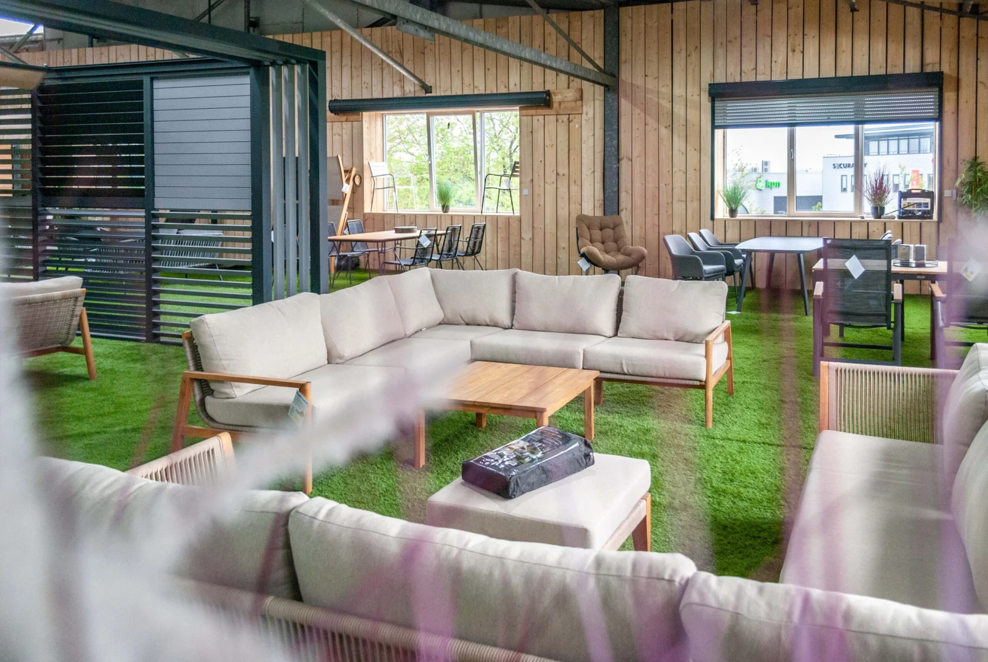Bestrating Zuid-holland Een ruime loungeset met beige kussens en houten frame, geplaatst in een overkapping met houten wanden en kunstgras. Op de achtergrond staan verschillende tuinmeubels, tafels en stoelen.
