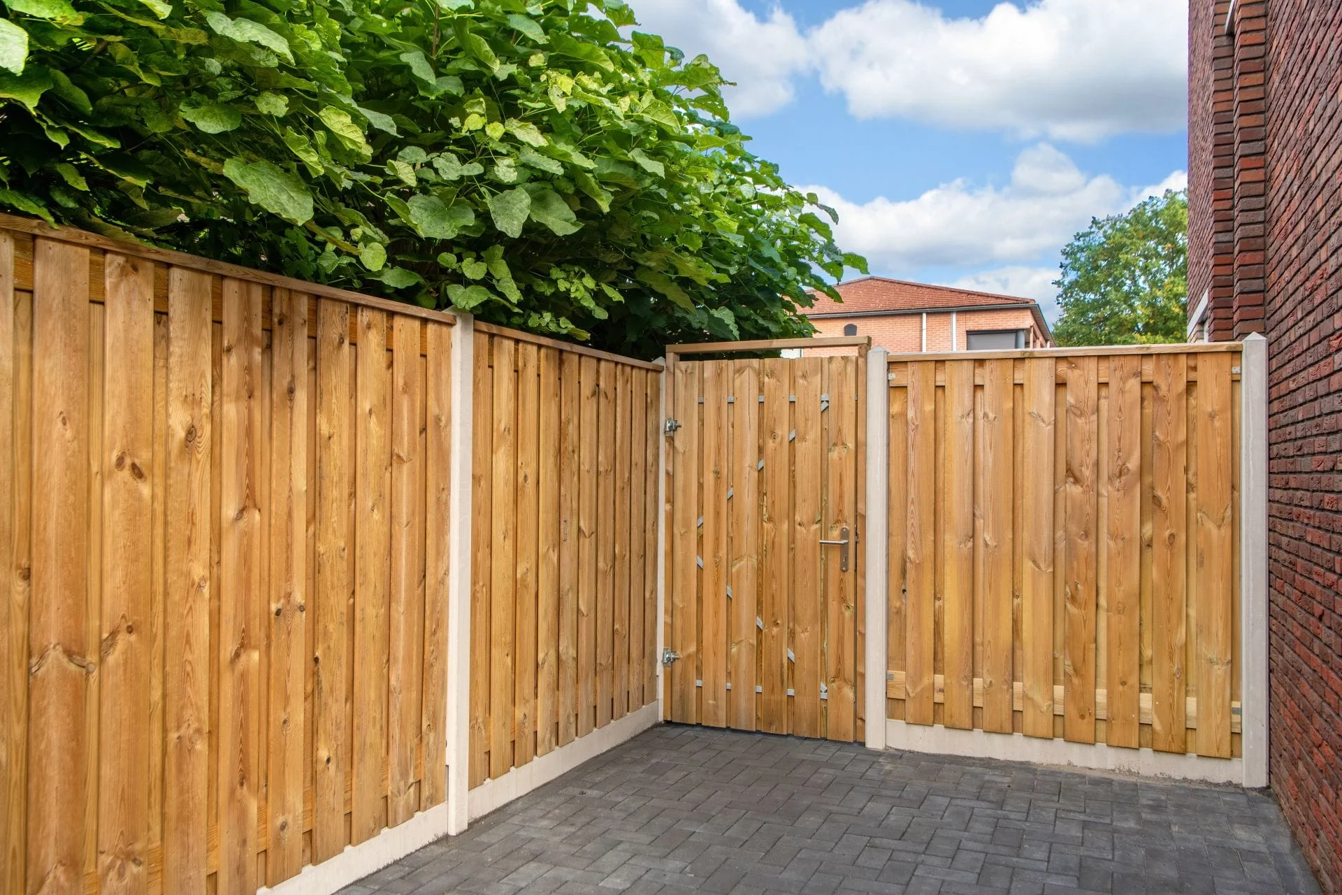 Betonpalen Een houten schutting met betonpalen en een bijpassende tuinpoort, geplaatst in een moderne tuin met bestrating en groene beplanting.