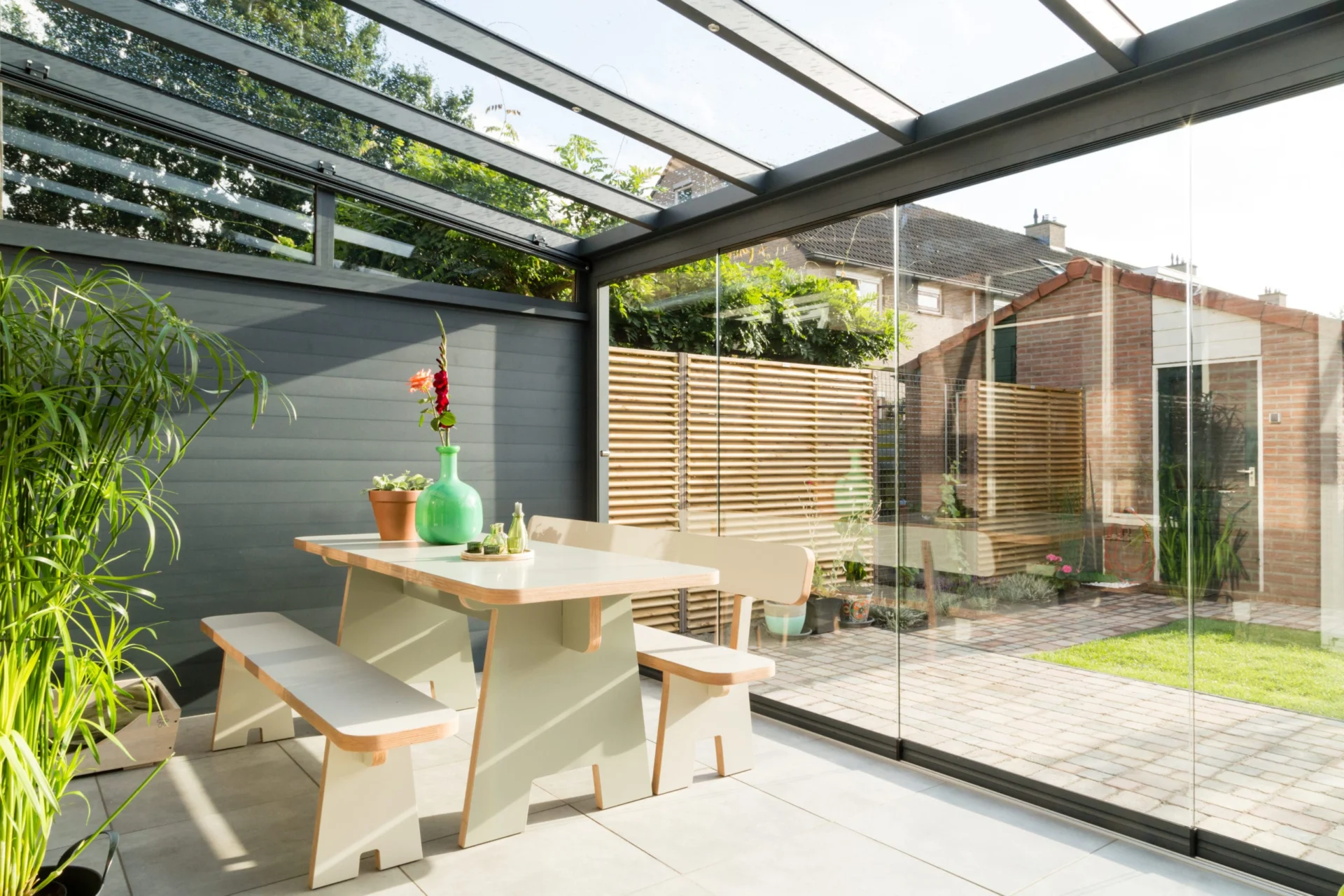 Tuinkamer Moderne Profiline tuinkamer in antraciet, voorzien van glazen schuifwanden voor een open en ruimtelijke uitstraling. De houten eethoek en groene accenten creëren een sfeervolle en natuurlijke ambiance.
