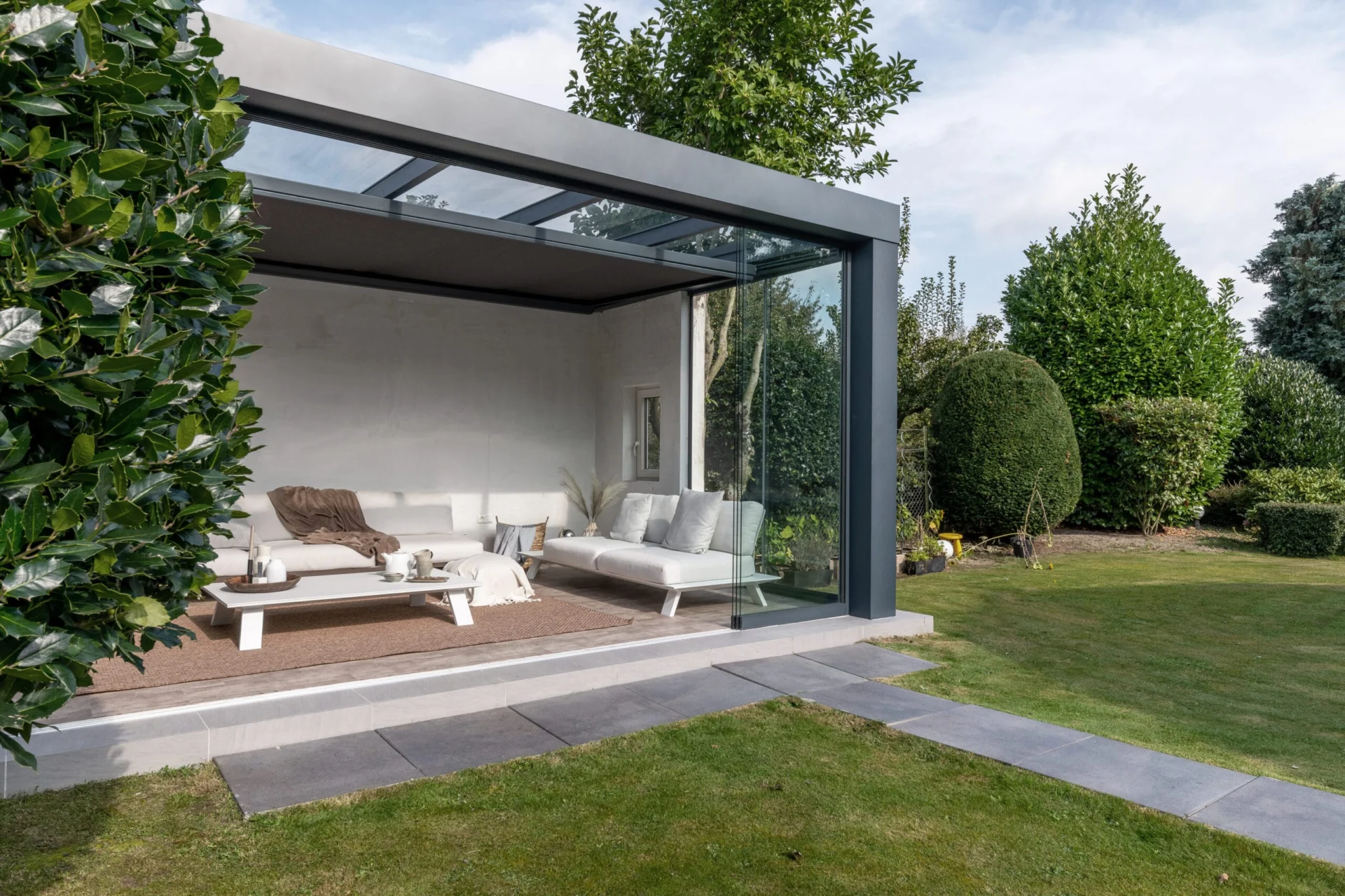 Tuinkamer Moderne Verasol Cube tuinkamer met een antraciet aluminium frame en glazen schuifwanden, ingericht als stijlvolle lounge in een groene tuin.