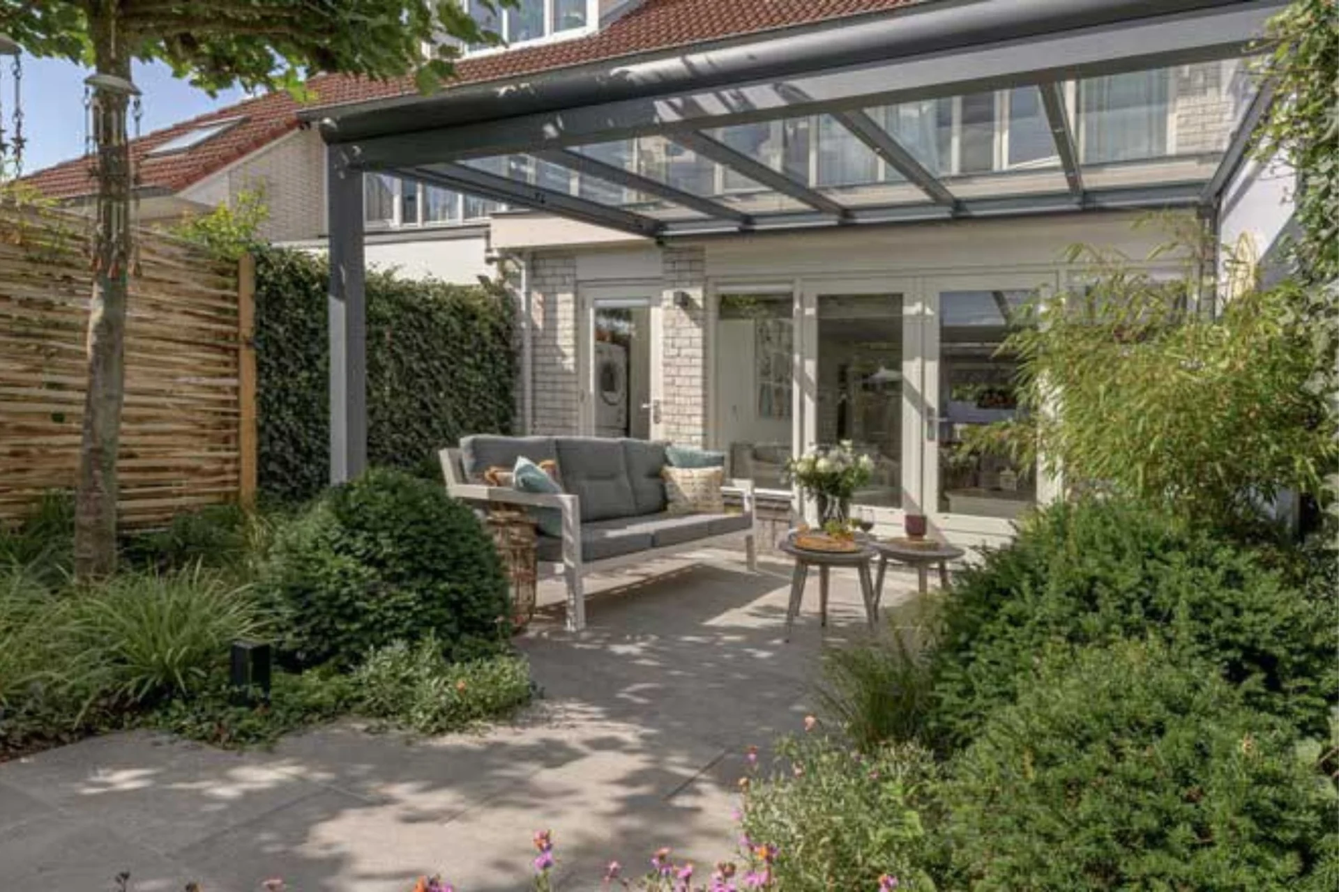 Veranda Luxe Greenline Verasol veranda met een stevig glazen dak, geplaatst in een sfeervolle tuin met een comfortabele loungeset en groene beplanting.