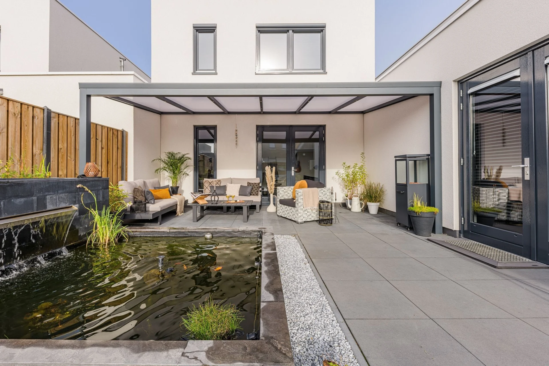 Veranda Moderne Greenline Verasol veranda met strakke antracietkleurige afwerking en een sfeervol ingerichte buitenruimte met loungeset en vijver.