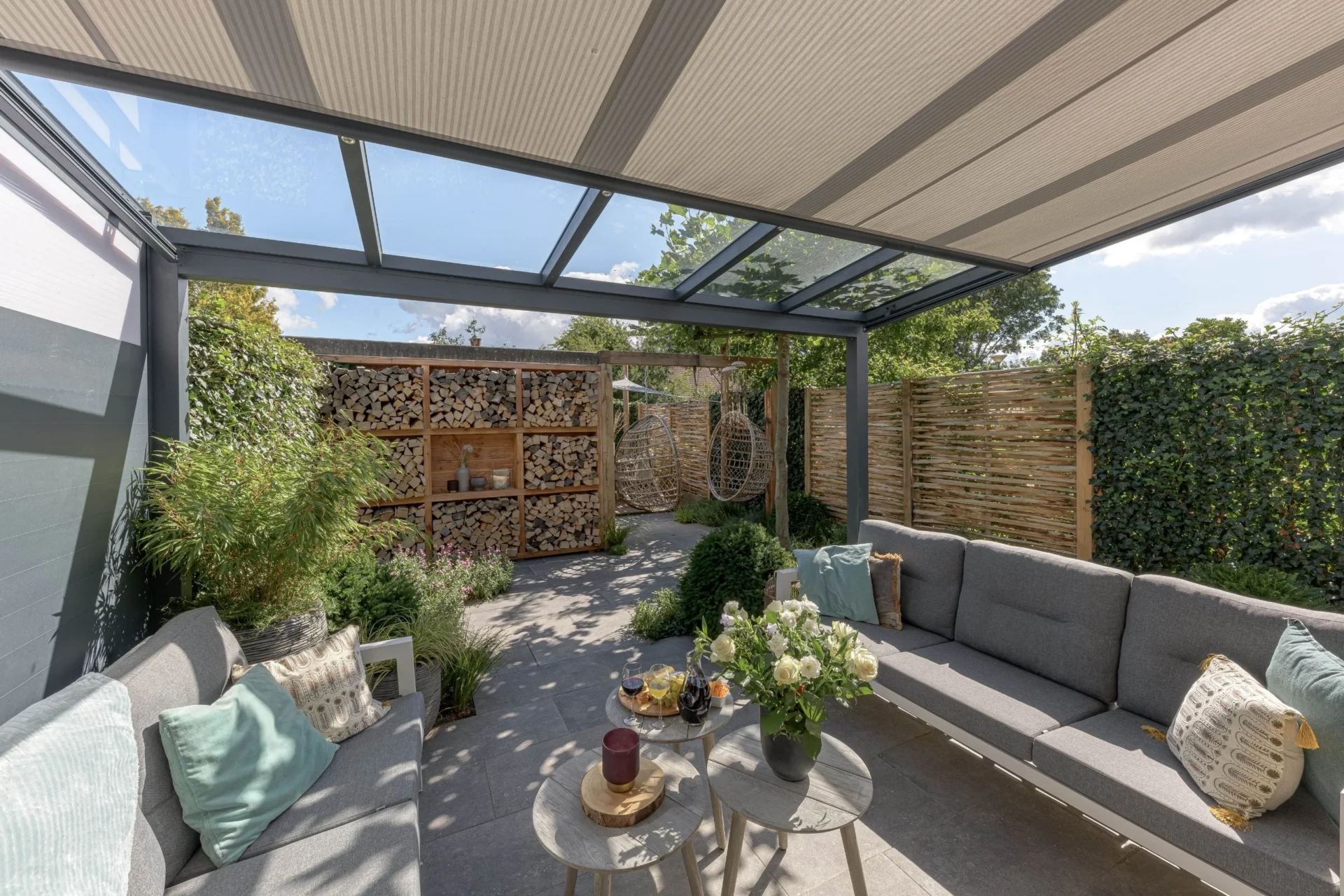 Veranda Gezellige buitenruimte met een Greenline Verasol veranda, geïntegreerde zonwering en een sfeervol ingerichte tuin met loungeset, haardhoutopslag en hangstoelen.