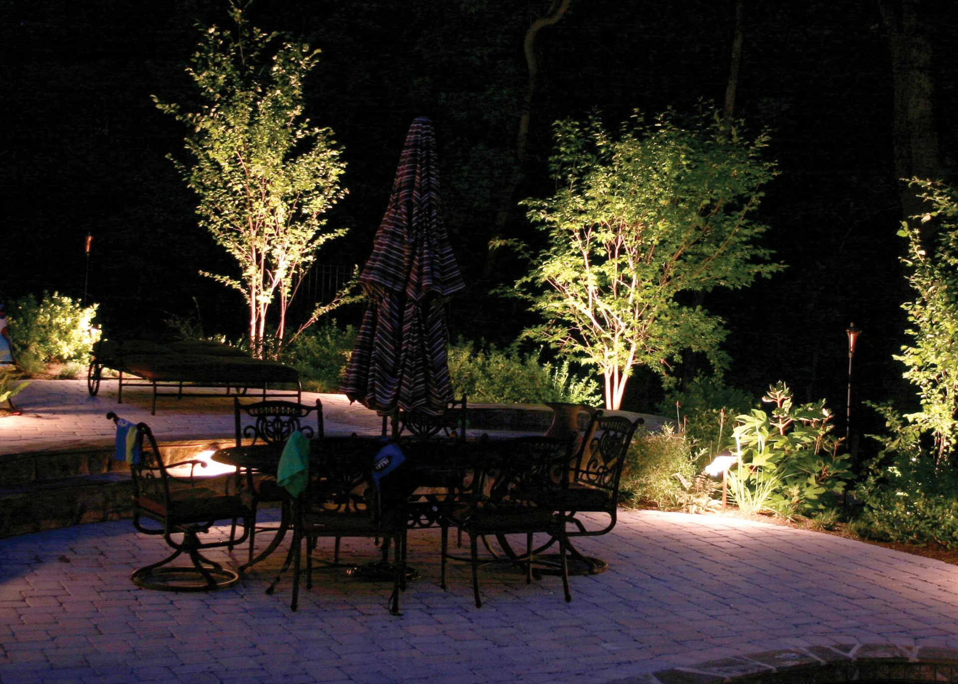 Bestrating Zuid-holland Verlichte tuin in de avond met sfeervolle spots die bomen, planten en een terras met metalen tuinmeubilair uitlichten.