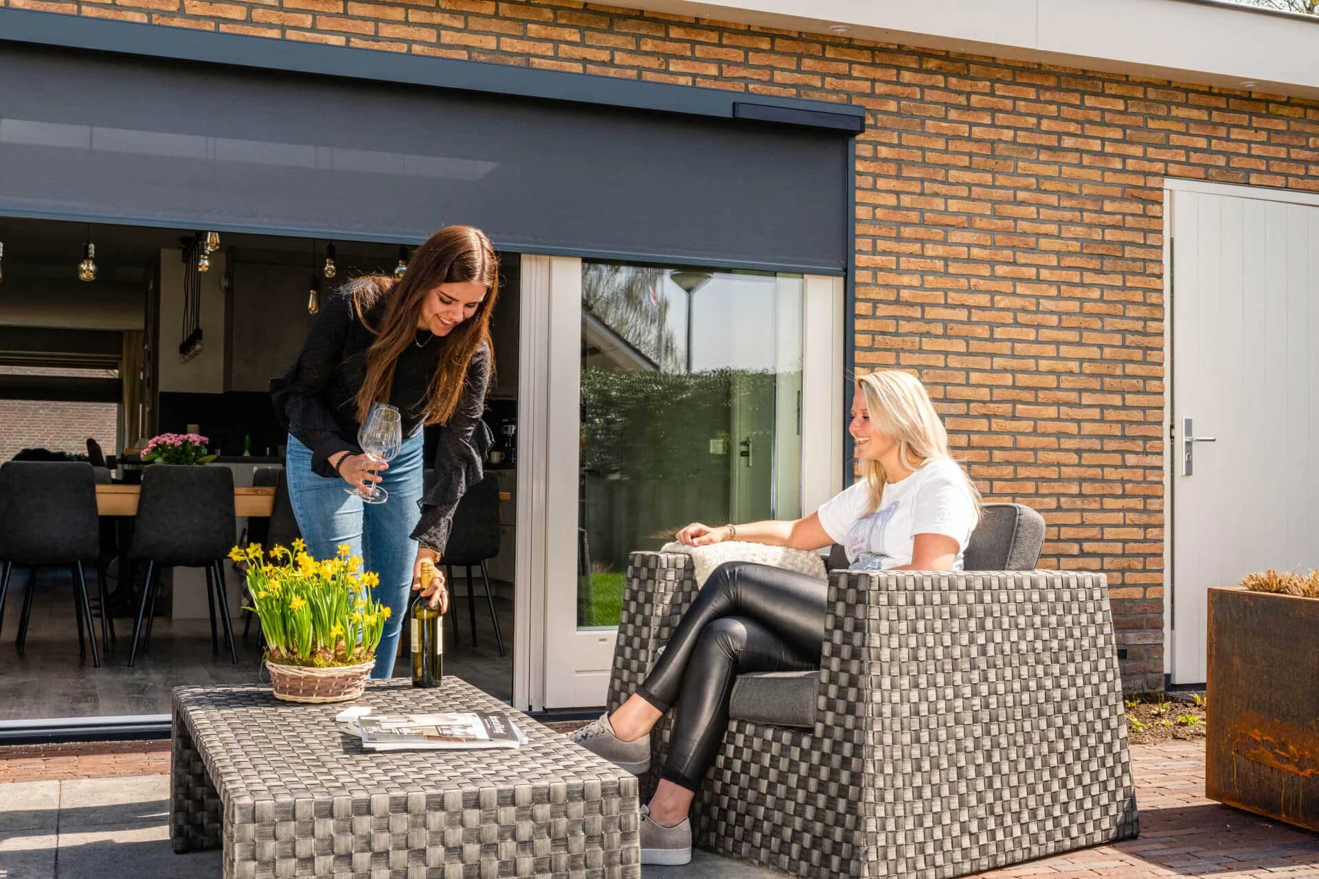 zonwering Donkergrijze screens gemonteerd op een bakstenen gevel, bieden effectieve zonwering en privacy voor een schuifpui met terras.