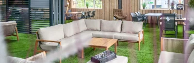 Een ruime loungeset met beige kussens en houten frame, geplaatst in een overkapping met houten wanden en kunstgras. Op de achtergrond staan verschillende tuinmeubels, tafels en stoelen.