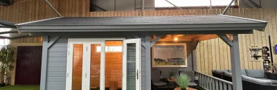 Een stijlvolle grijze blokhut met witte deuren en een aangebouwde overkapping, tentoongesteld in de showroom van Livingstone Goes. De blokhut is voorzien van een gezellige zithoek en kunstgras als ondergrond.