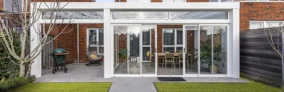Een moderne witte overkapping met glazen schuifwanden, aangebouwd aan een rode bakstenen woning. De overkapping heeft een luxe inrichting met een houten eettafel, stoelen en planten, en grenst aan een tuin met kunstgras en grijze tuintegels.